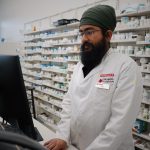 Farmacêutico trabalha diante de um computador em uma farmácia com prateleiras de medicamentos ao fundo.