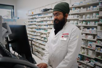 Farmacêutico trabalha diante de um computador em uma farmácia com prateleiras de medicamentos ao fundo.
