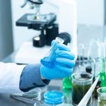 Cientista usando luvas analisa um líquido azul em um laboratório, ao lado de um microscópio e vidrarias.