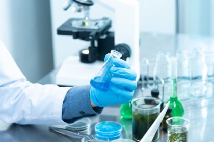 Cientista usando luvas analisa um líquido azul em um laboratório, ao lado de um microscópio e vidrarias.